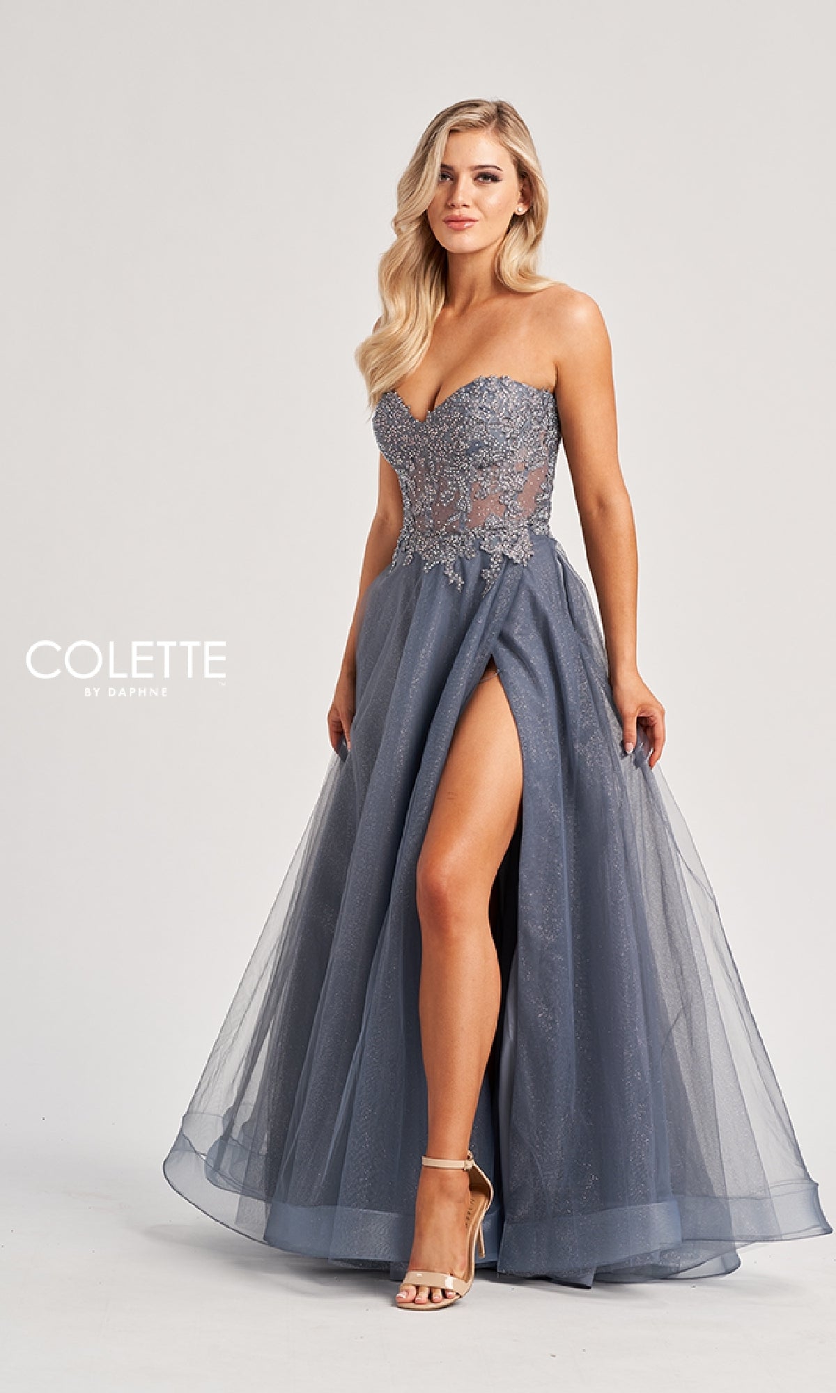 A woman poses elegantly in the Long Prom Dress: Colette CL8110, a strapless ball gown with an embroidered corset bodice, sheer detailing, and a high slit, set against a plain background.